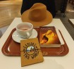 Na zdjęciu stół z książką, filiżanką herbaty, kapeluszem i notesem.
